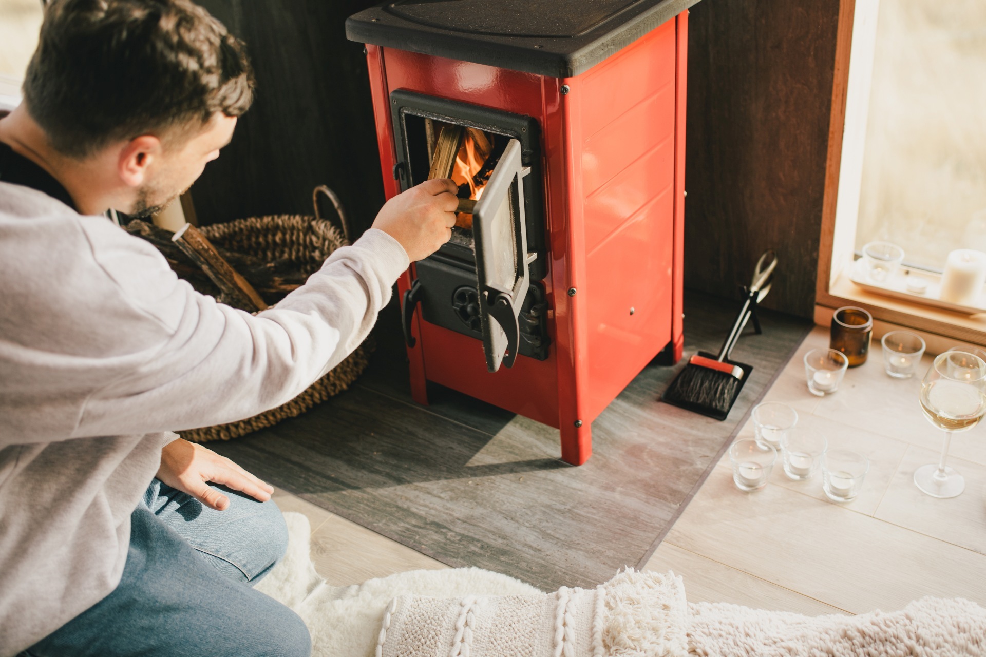 The photo shows a man kneeling in front of a wood-burning stove and putting a log into the fire through the open stove door.