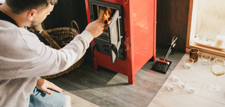 The photo shows a man kneeling in front of a wood-burning stove and putting a log into the fire through the open stove door.