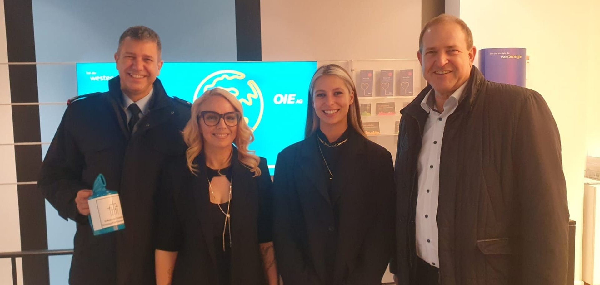 The photo shows the people mentioned. They are standing next to each other in front of the advertising wall and looking into the camera. Colonel Tuneke is holding a donation box in his hand.