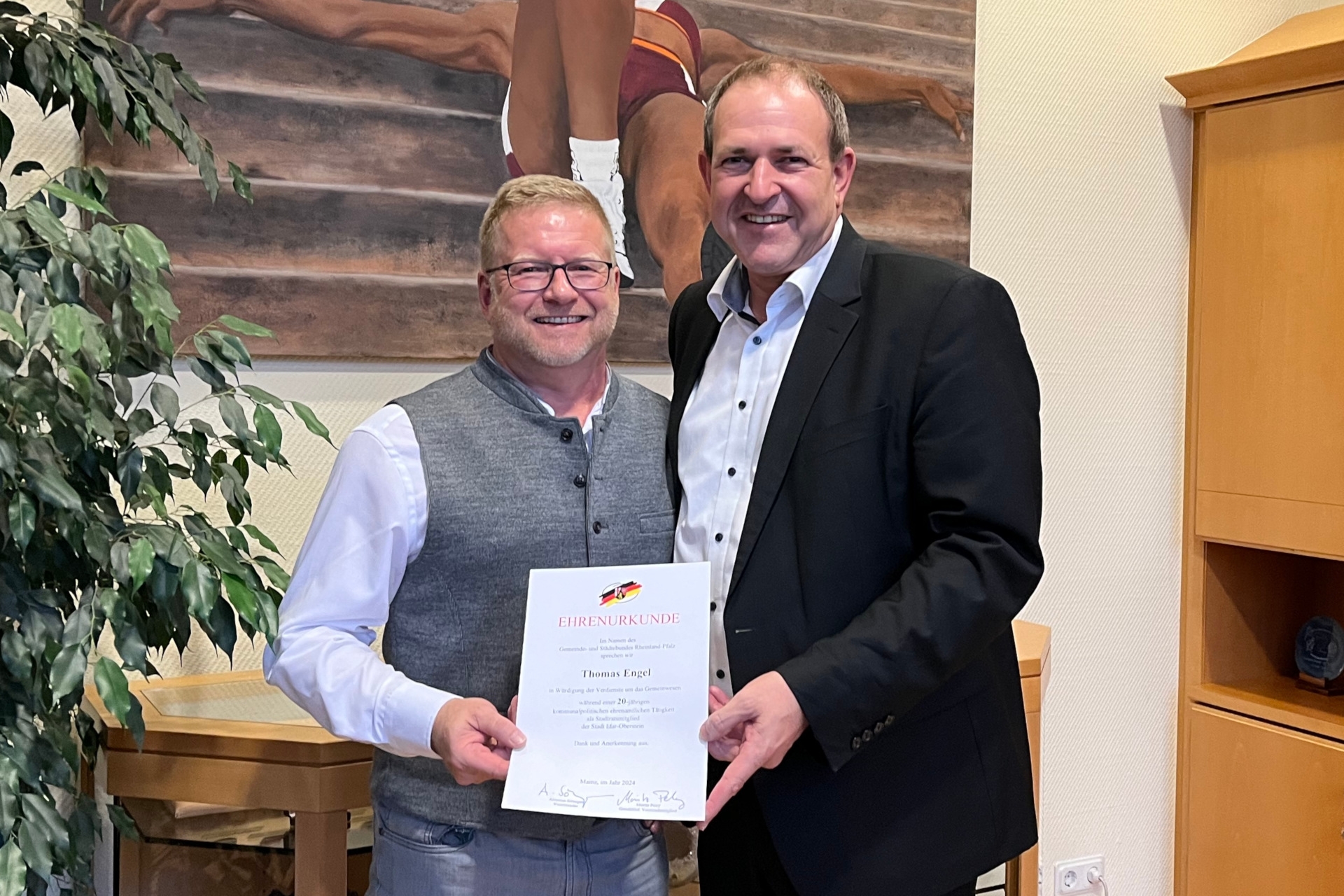 The photo shows the two people. They are standing next to each other in the Lord Mayor's office, holding a certificate together and looking into the camera.