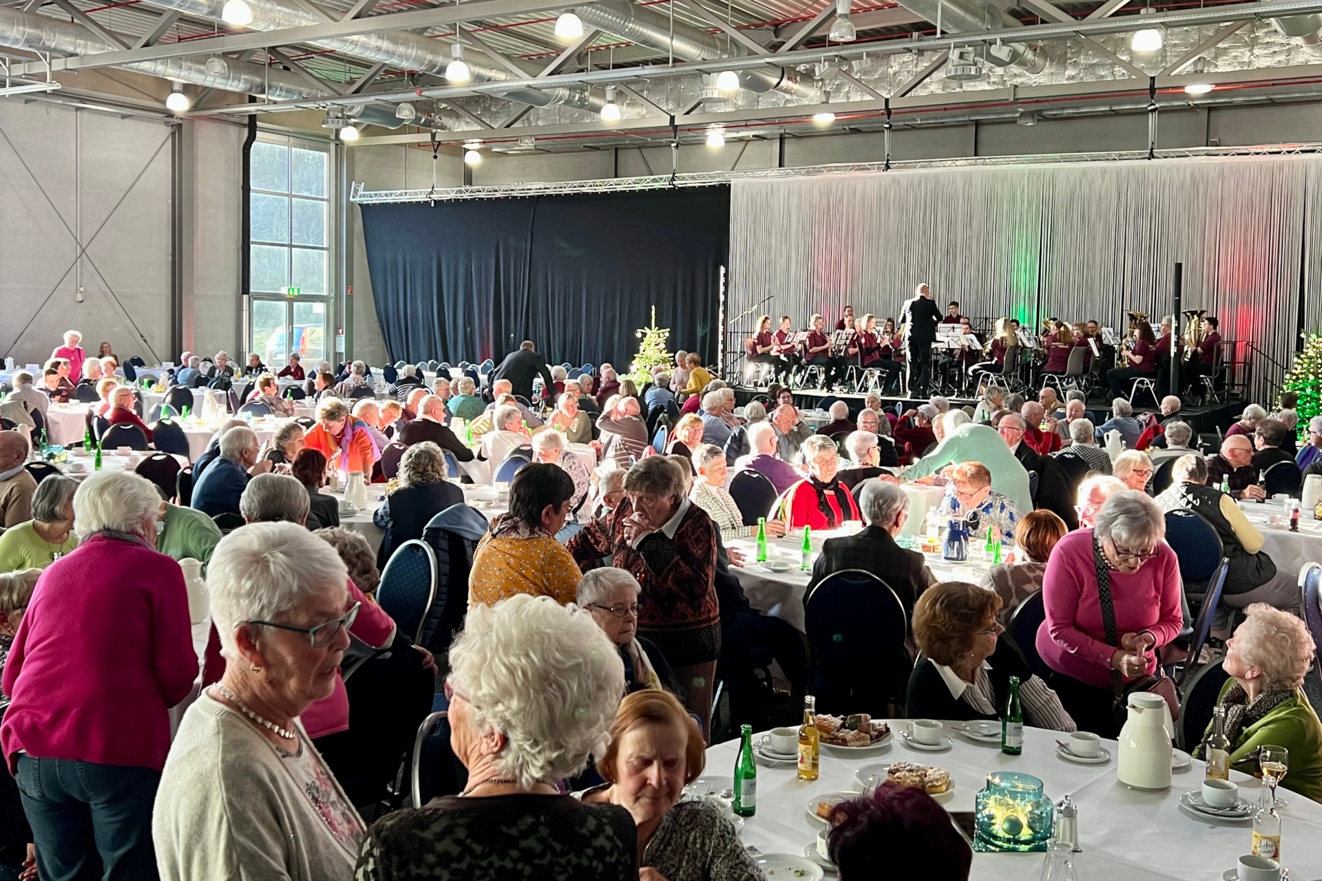 The photo shows a view of the well-filled exhibition hall. The guests are seated at round tables, the stage can be seen in the background. The Tiefenstein Music Society is playing there.