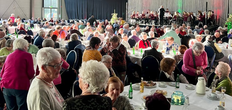 The photo shows a view of the well-filled exhibition hall. The guests are seated at round tables, the stage can be seen in the background. The Tiefenstein Music Society is playing there.