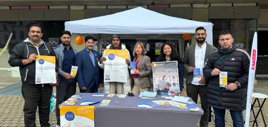The photo shows the information stand with flyers and posters. Several people are standing at the stand, also holding flyers and posters and looking into the camera.