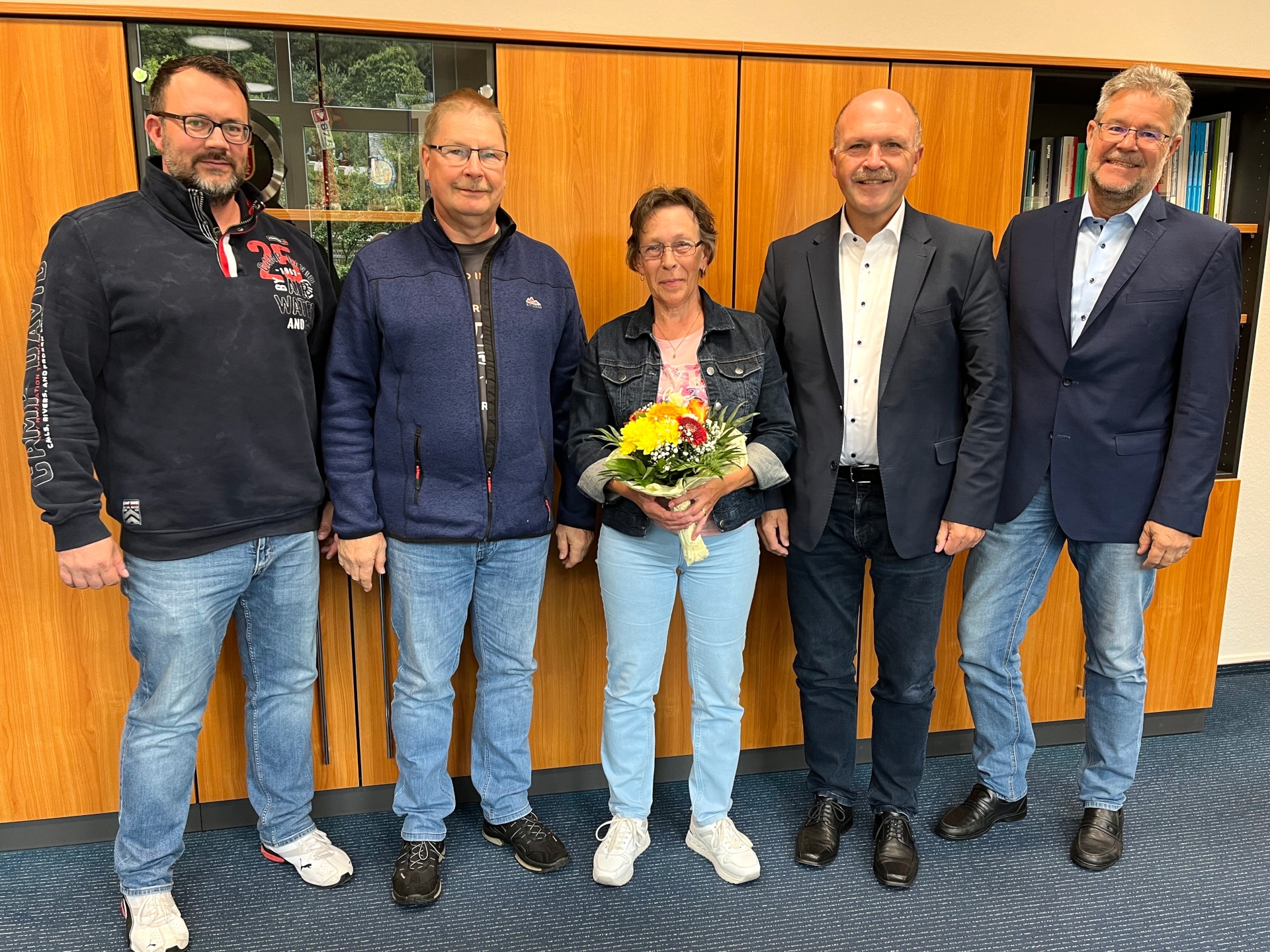 The photo shows five people standing next to each other in front of a wall unit and looking into the camera. Mrs. Di Cato stands in the middle with a bouquet of flowers in her hands.