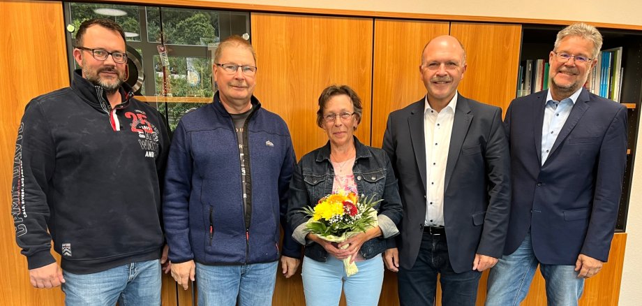 The photo shows five people standing next to each other in front of a wall unit and looking into the camera. Mrs. Di Cato stands in the middle with a bouquet of flowers in her hands.