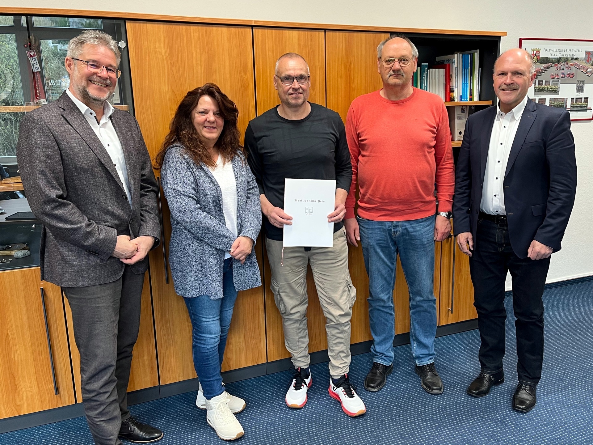 The photo shows the five participants in the ceremony. They are standing next to each other in front of a wall unit in Mayor Marx's office and looking into the camera. Rolf Zehmke holds the certificate of thanks in his hands.