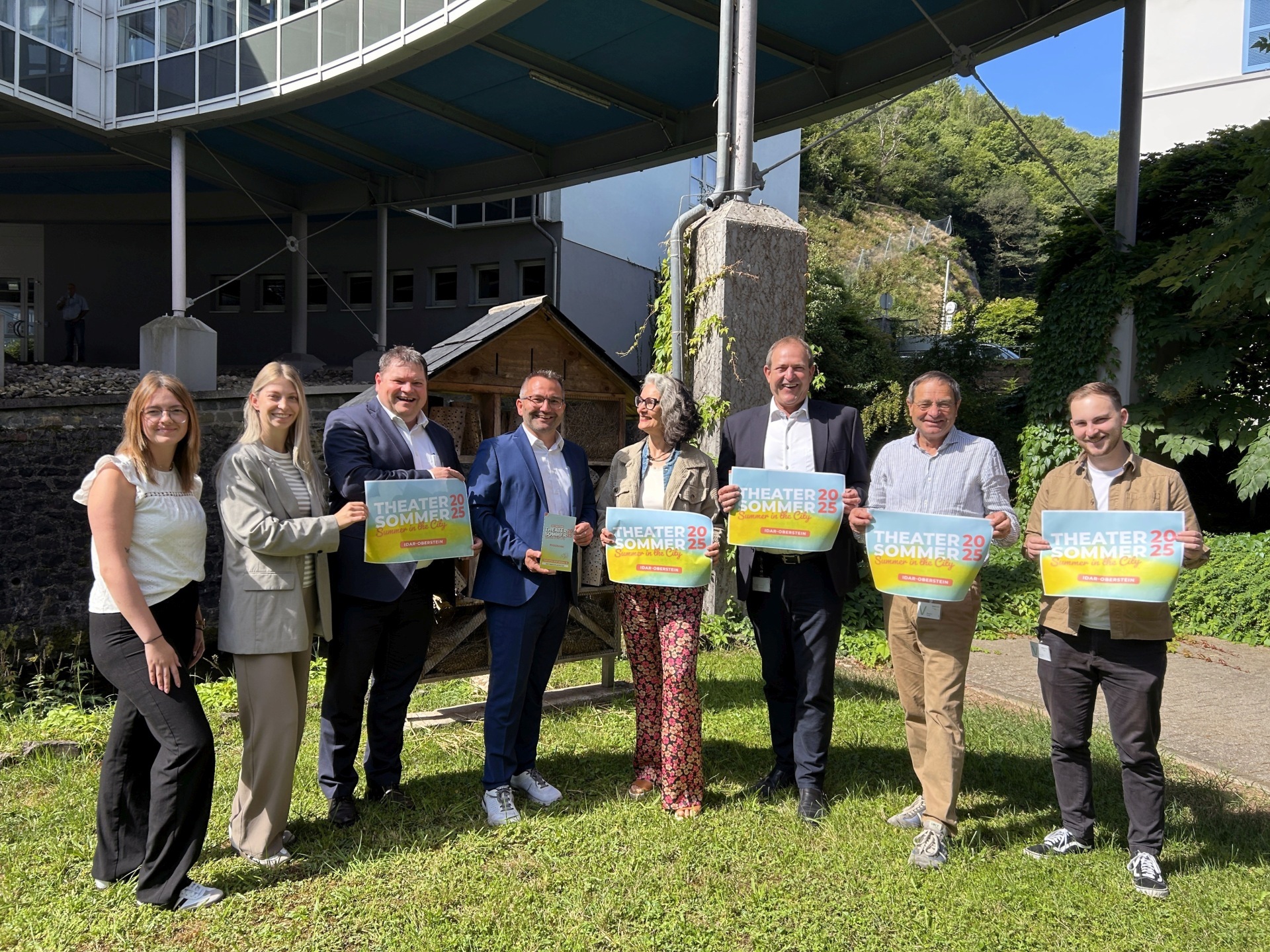The photo shows the organizers and sponsors. They are standing next to each other in the garden of the OIE building, holding posters for the theater summer.