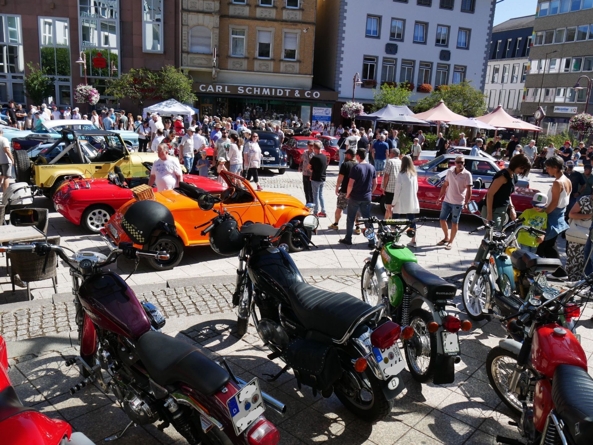 The photo shows the Idar Schleiferplatz with many vintage cars and visitors. Historic motorcycles are on the stage of the concert shell.