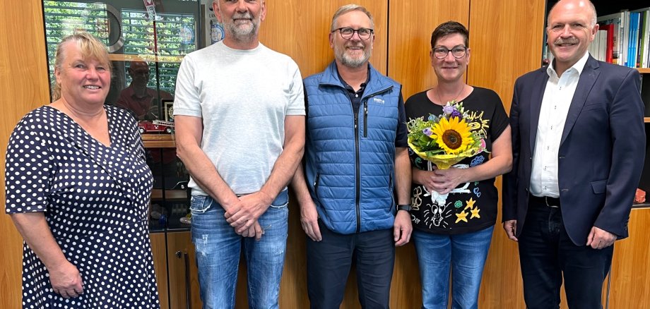 The photo shows the aforementioned people standing next to each other in front of a wall unit and looking into the camera. Nicole Hermann holds a bouquet of flowers in her hands.