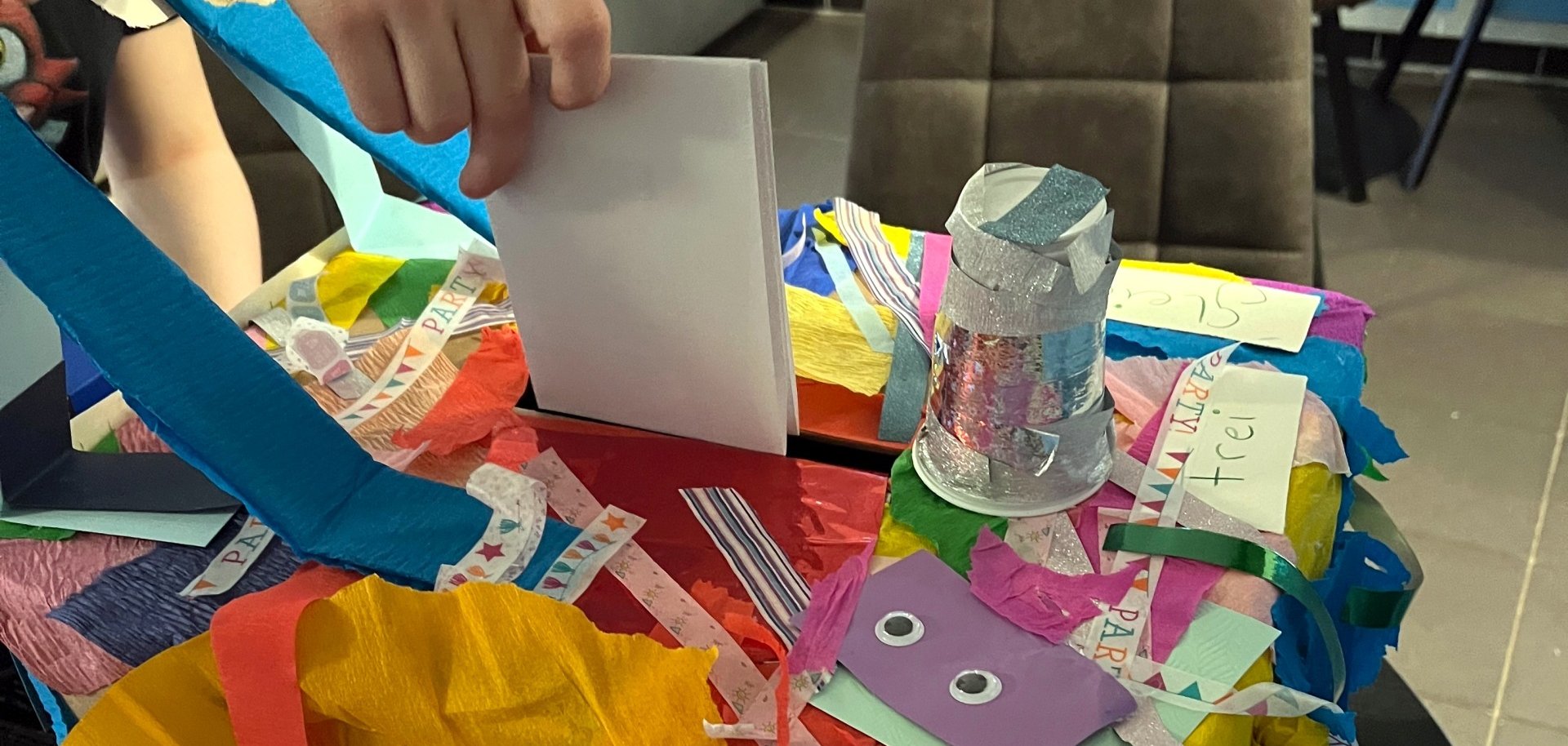 The photo shows a hand dropping a ballot paper into a colorfully decorated ballot box.
