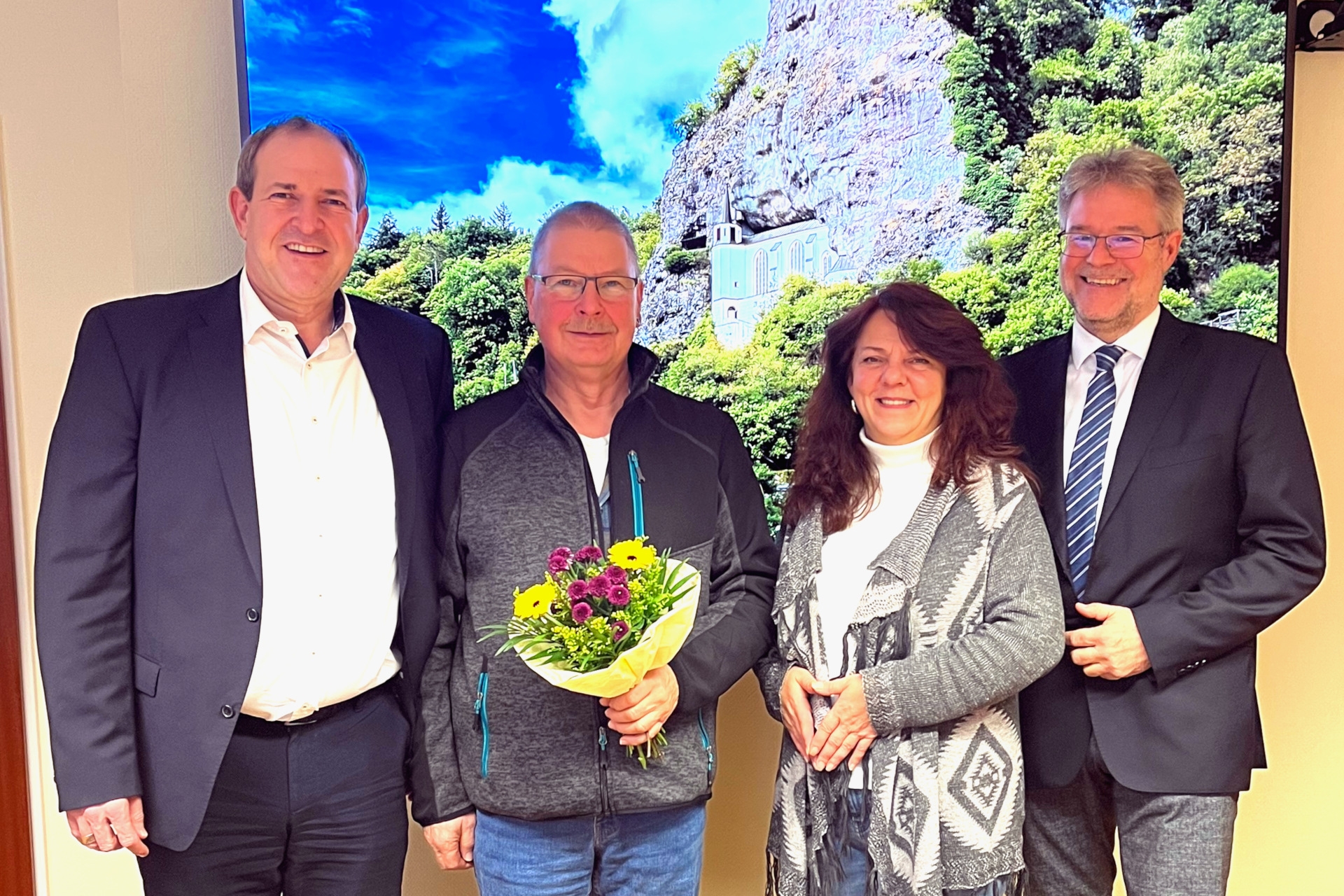 The photo shows the people mentioned. They are standing next to each other in front of a picture of the Felsenkirche and looking into the camera. Volker Poeß is holding a bouquet of flowers.