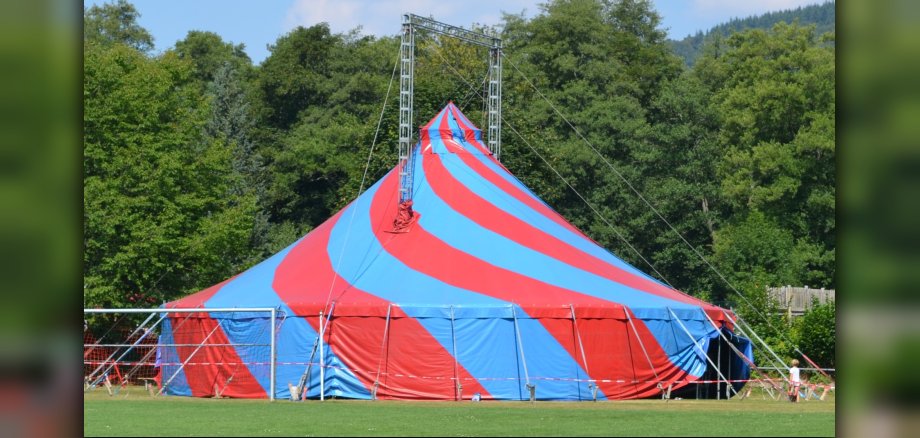 The photo shows a large, blue and red striped circus tent standing on a meadow.