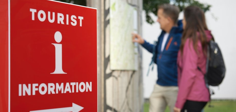 The photo shows the i-mark in the foreground and hikers in front of a map in the background.