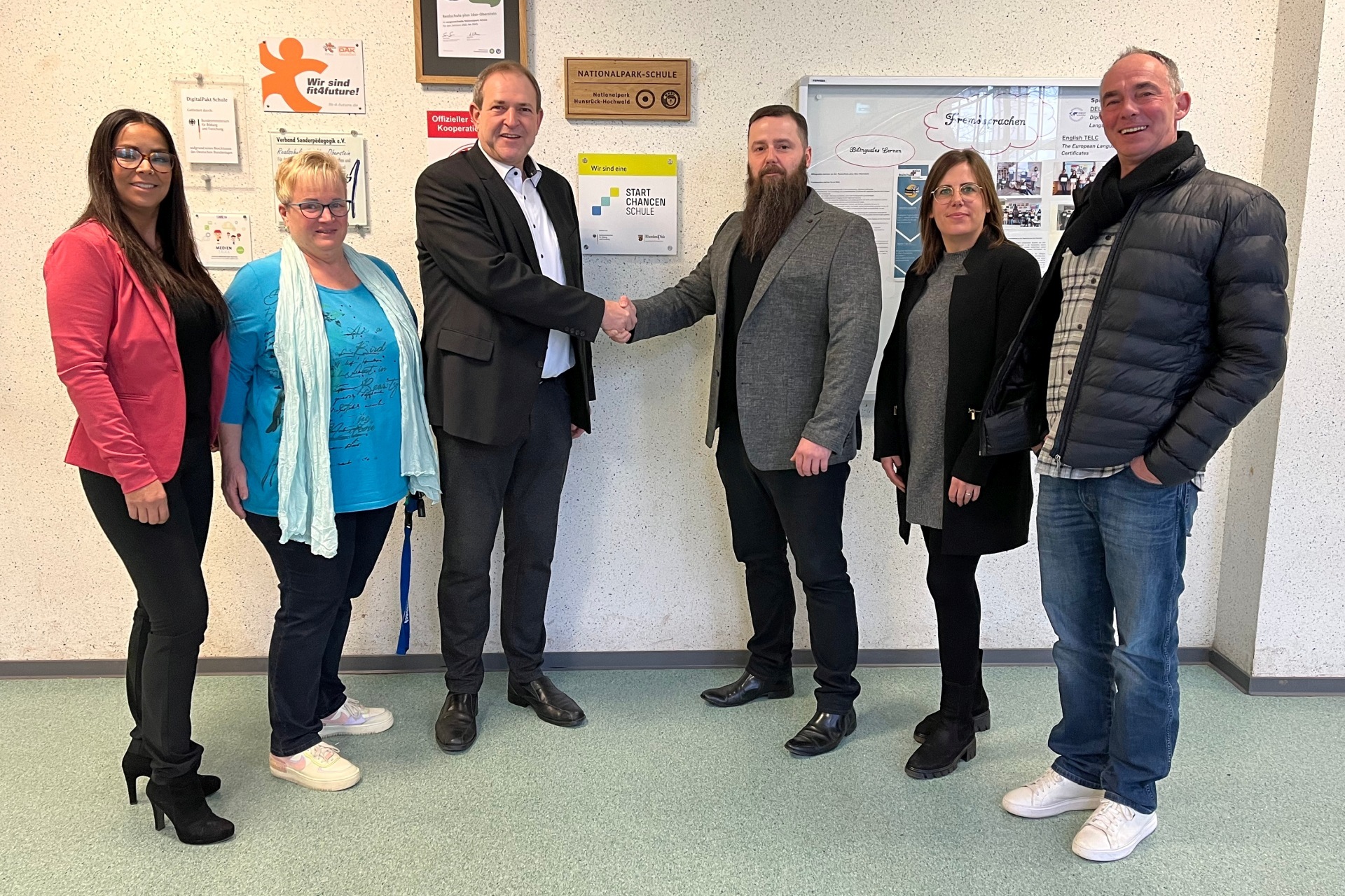 The photo shows the people mentioned. They are standing next to each other in front of the wall on which the Startchancen program plaque is attached. Mayor Frühauf and principal Back shake hands.