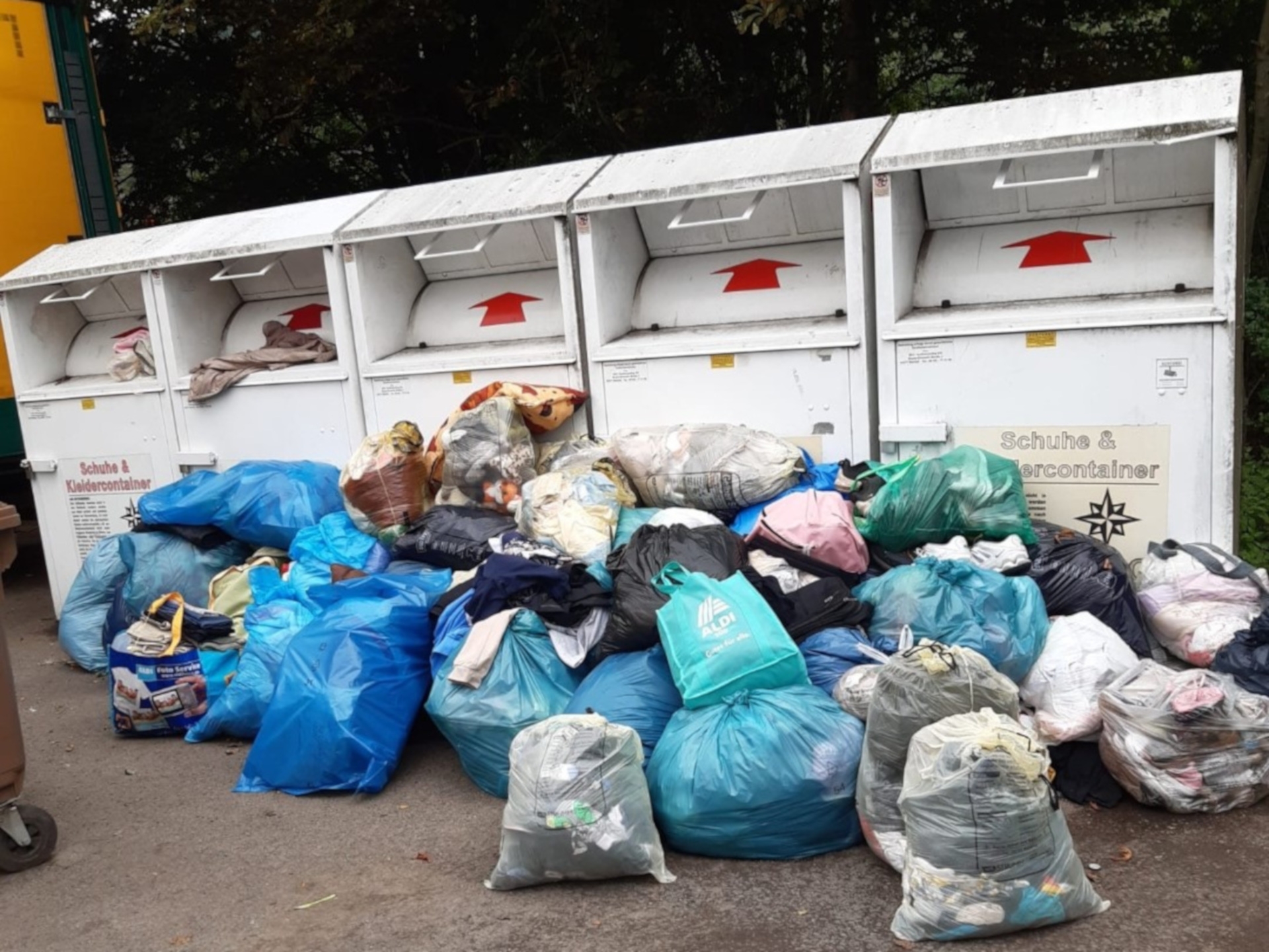 The photo shows old clothes containers in front of which numerous plastic bags with more old clothes are lying.