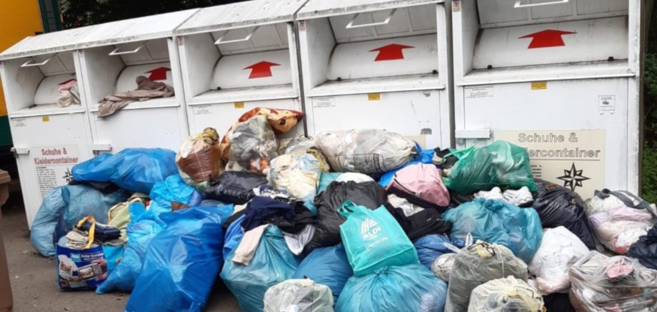 The photo shows old clothes containers in front of which numerous plastic bags with more old clothes are lying.