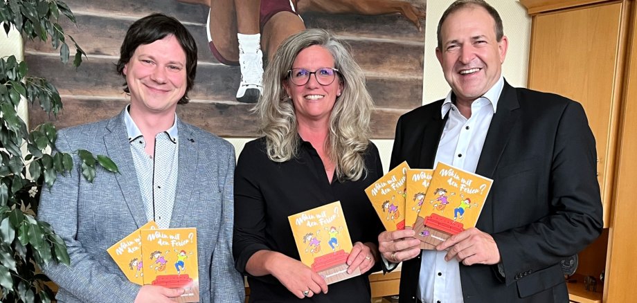 The photo shows the people mentioned. They are standing next to each other in the mayor's office, looking into the camera and each holding a vacation booklet.