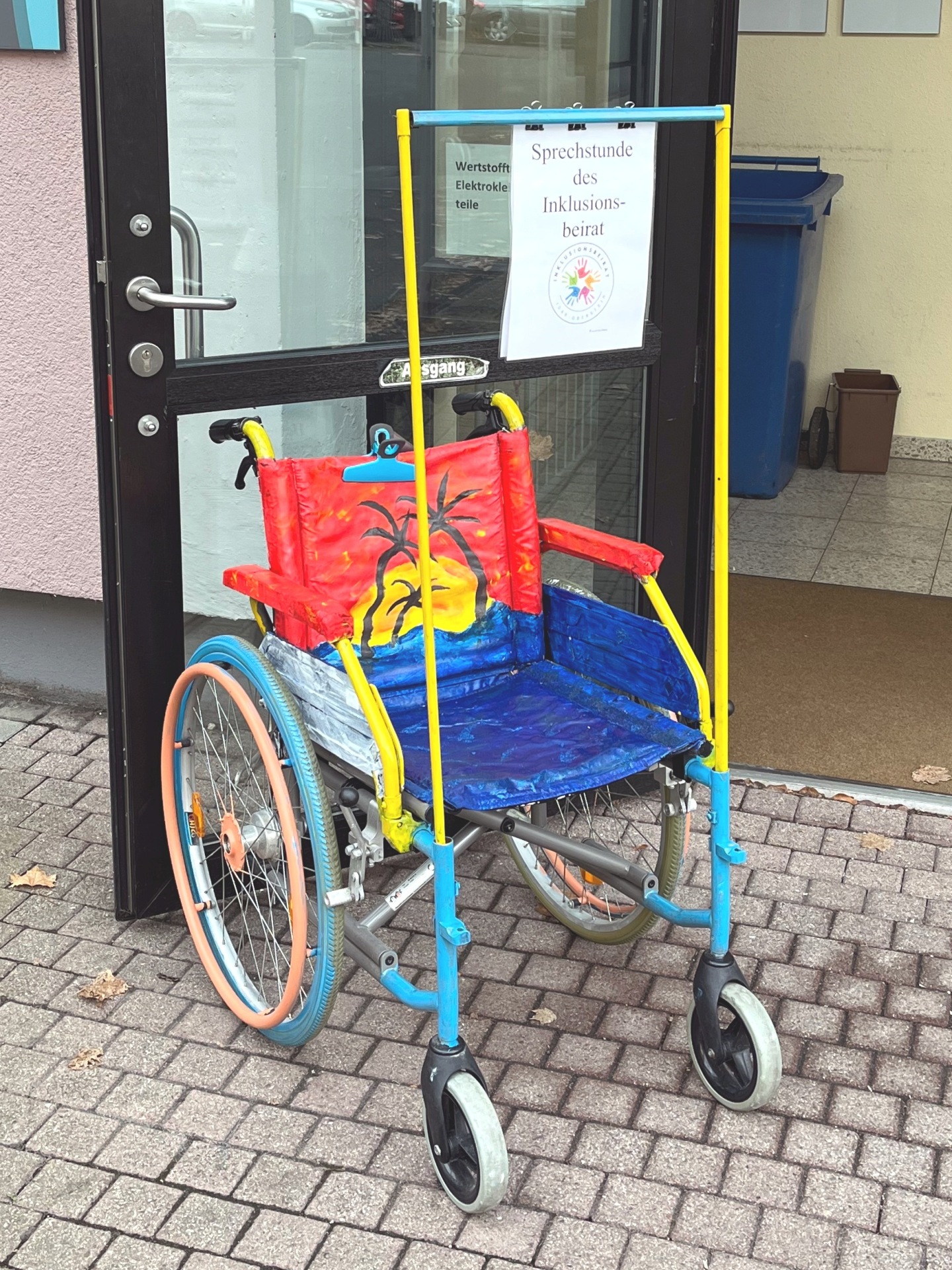 The photo shows the colorful wheelchair in front of the "glass box" where the consultation hours of the Inclusion Advisory Board take place.