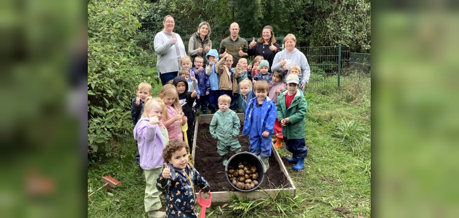 The Enzweiler daycare center proudly presents its fantastic potato harvest. The photo shows teachers, janitors and children standing around the planting bed and presenting a bucket full of harvested potatoes.