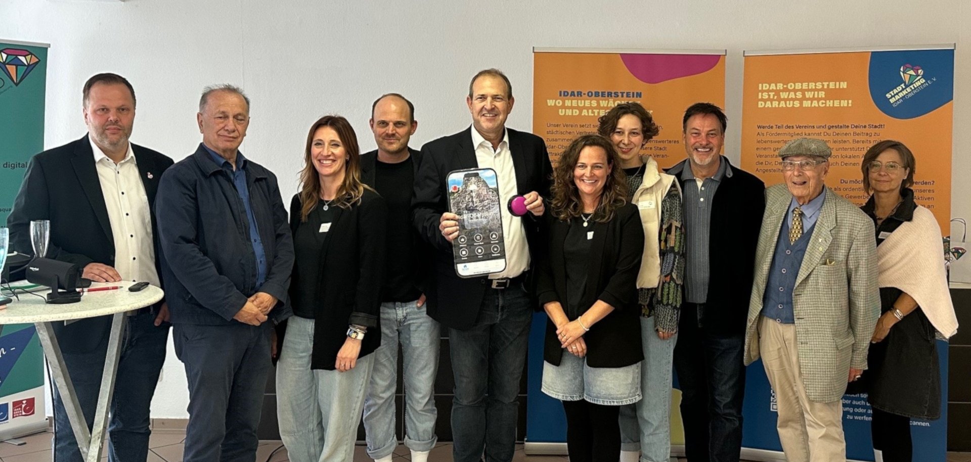 The photo shows several people in front of roll-ups of the support program. Mayor Frühauf is holding a button and a graphic of the app.