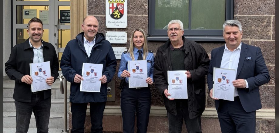 First Chief Detective Inspector Timo Jung, Mayor Friedrich Marx, Police Councillor Kimberly Short, Association Mayor Uwe Weber and District Administrator Miroslaw Kowalski present the signed security partnership. The photo shows the people mentioned. They are standing in front of the police station building and holding the documents up to the camera.