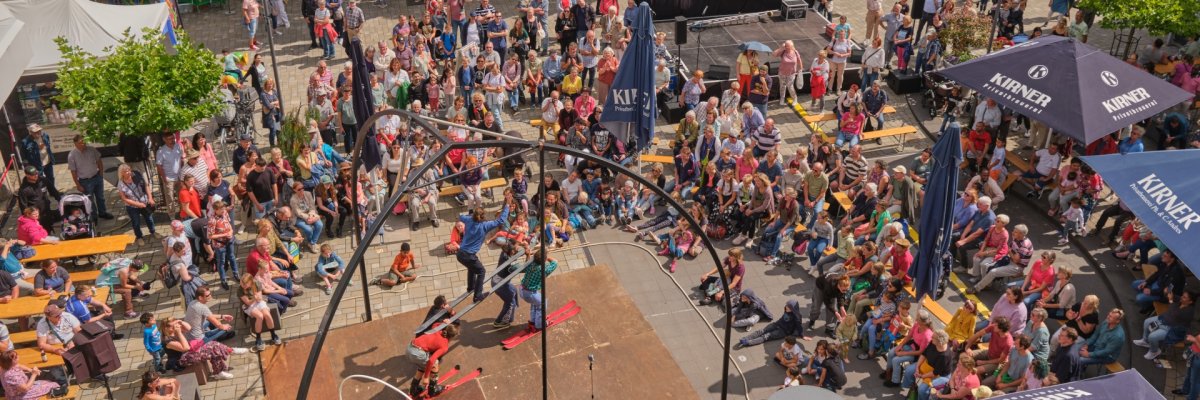 Festival de théâtre de rue 2023 La photo montre une vue d'en haut de la scène de la place de la Christuskirch. Sur la scène, on peut voir des artistes sur des skis, devant lesquels un grand nombre de spectateurs sont assis.