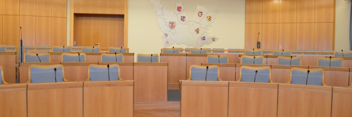 Salle de réunion de la municipalité d'Idar-Oberstein La photo montre une vue de la salle de réunion. Les rangées de chaises des membres du conseil sont disposées en demi-cercle de manière ascendante. Sur le mur derrière, une installation montre les contours de la ville d'Idar-Oberstein avec les armoiries des différents quartiers.