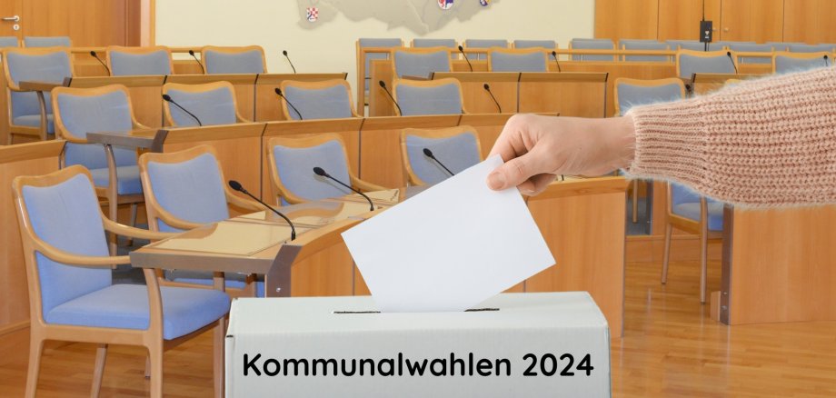 La photo montre un bras qui met un bulletin de vote dans une urne. En arrière-plan, on peut voir la salle de réunion de la municipalité d'Idar-Oberstein.