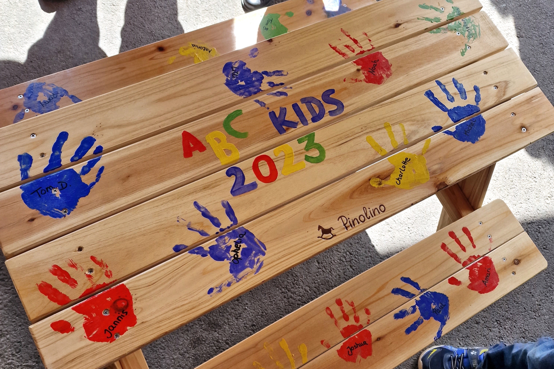 La photo montre un ensemble de sièges en bois avec une table et deux bancs. Sur la garniture, on peut voir des empreintes de mains colorées des enfants ABC avec le nom de chacun d'entre eux.