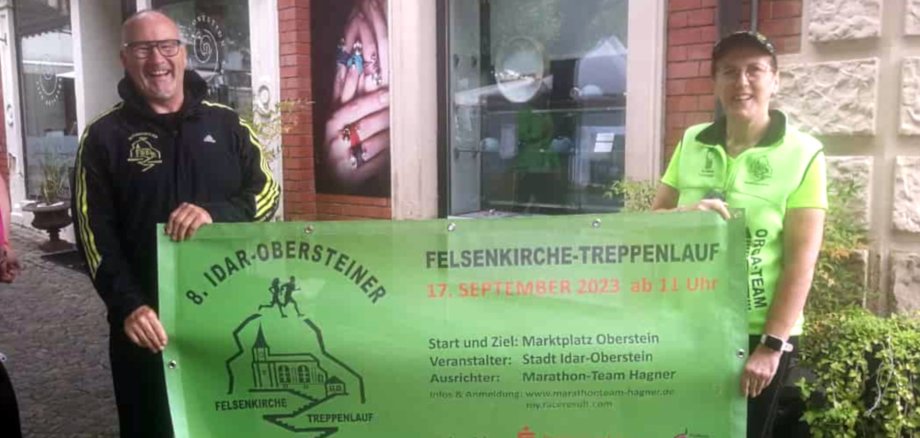 La photo montre le couple Rainer et Ilonka Hagner. Ils tiennent une bannière publicitaire pour la 8e édition de la Felsenkirche-Treppenlauf d'Idar-Oberstein.