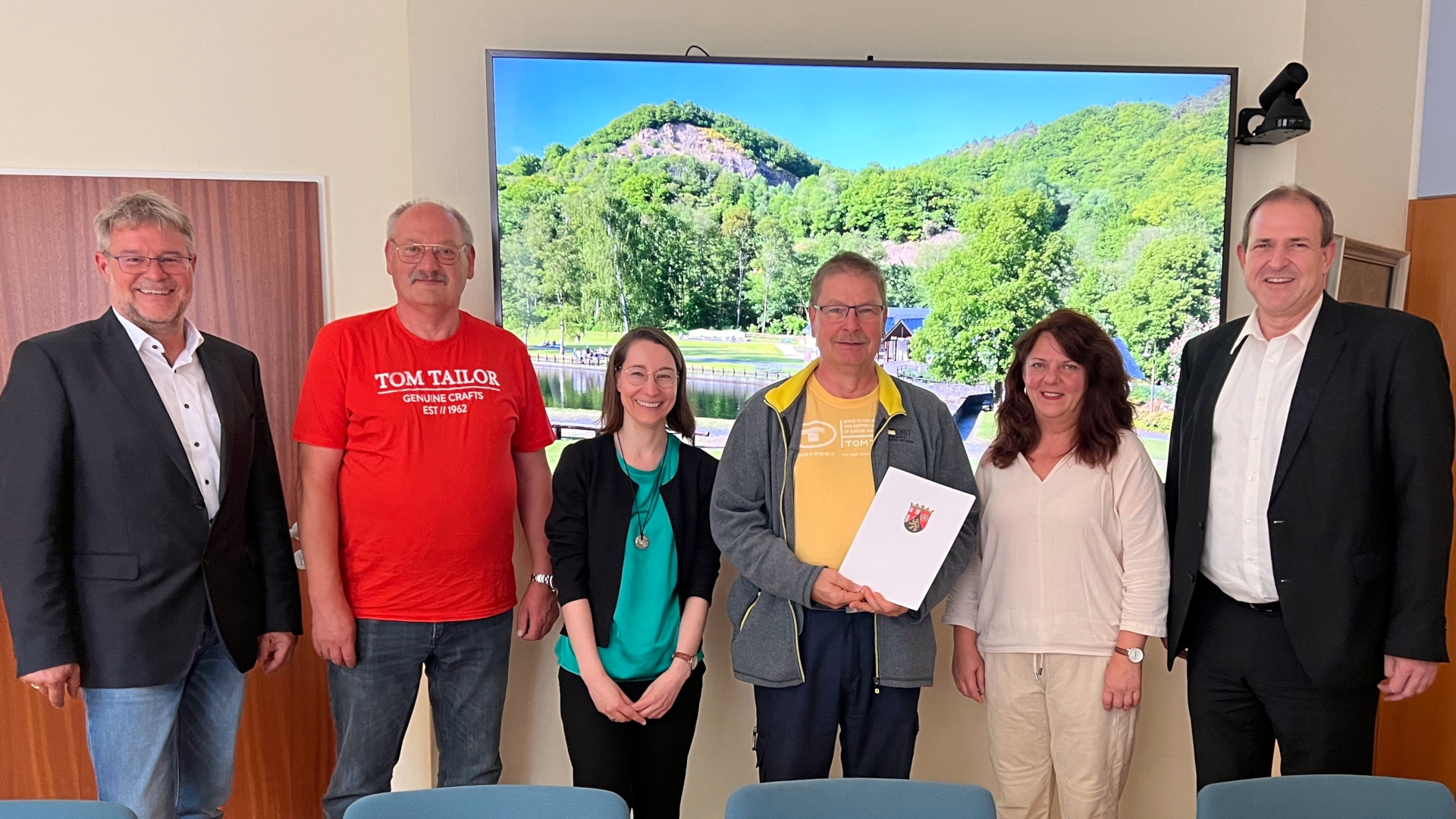 La photo montre les personnes mentionnées, debout côte à côte. Volker Poes tient le certificat de la Première ministre dans sa main.