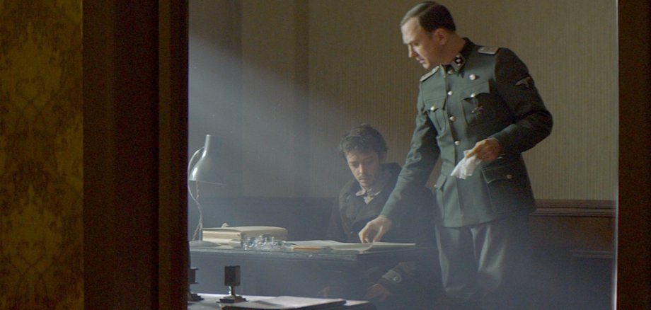 La photo montre deux hommes dans une pièce sombre. L'un est assis à un bureau. L'autre se tient à côté de lui et feuillette un livre posé sur le bureau.