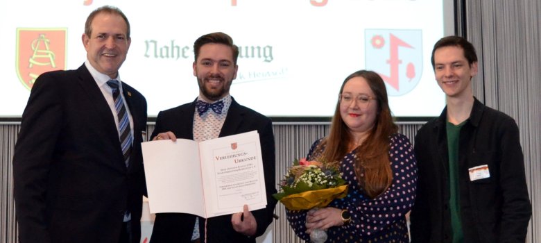 La photo montre les personnes mentionnées. Elles se tiennent côte à côte sur la scène et regardent la caméra. Moritz Forster présente le certificat de nomination.