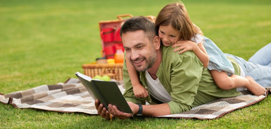 La photo montre un homme allongé sur une pelouse en train de lire un livre. Une fillette est allongée sur son dos et participe à la lecture.