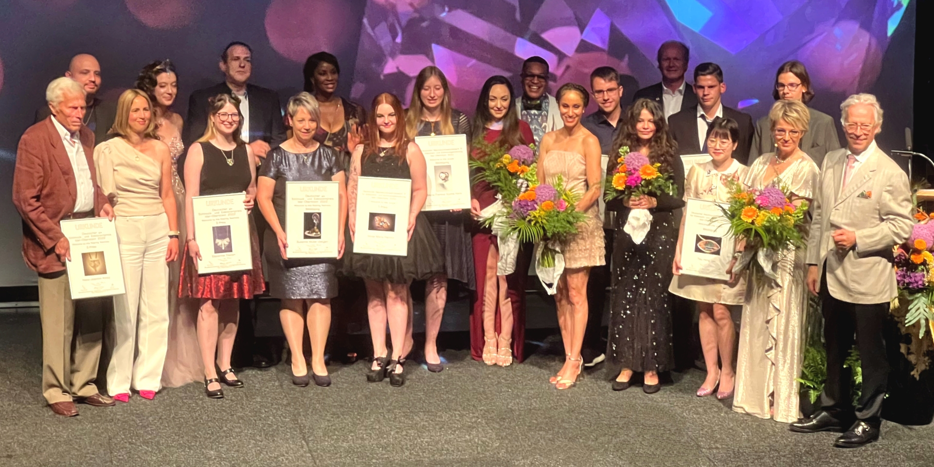 La photo montre de nombreuses personnes sur la scène du théâtre municipal : les organisateurs, la présentatrice les artistes et les lauréats avec leurs diplômes,