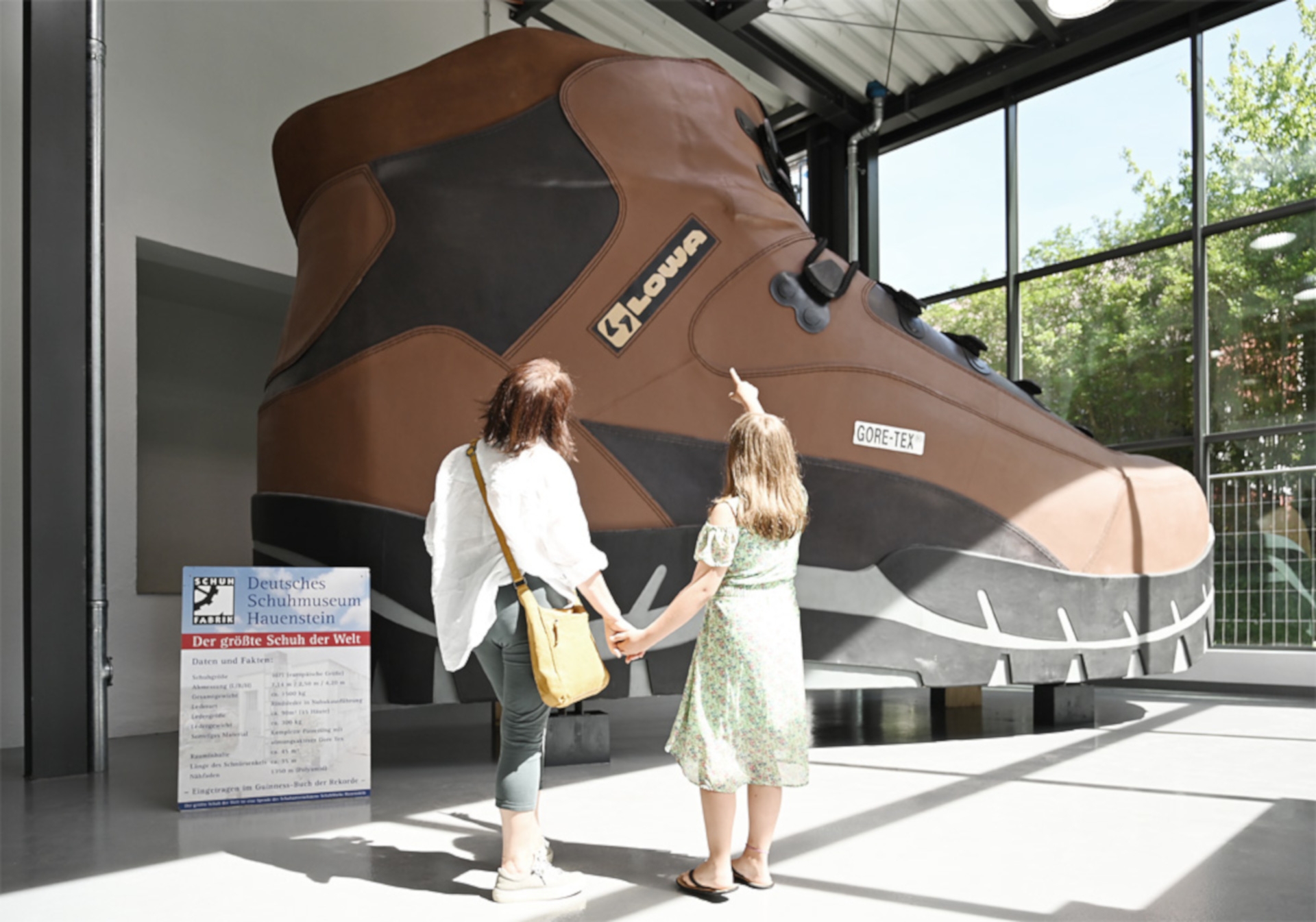La photo montre deux visiteurs se tenant devant la plus grande chaussure du monde devant le musée de la chaussure Hauenstein.
