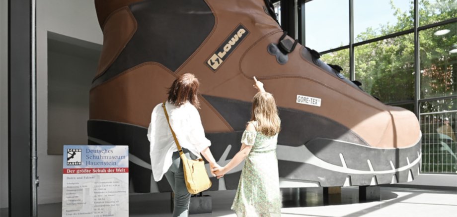 La photo montre deux visiteurs se tenant devant la plus grande chaussure du monde devant le musée de la chaussure Hauenstein.