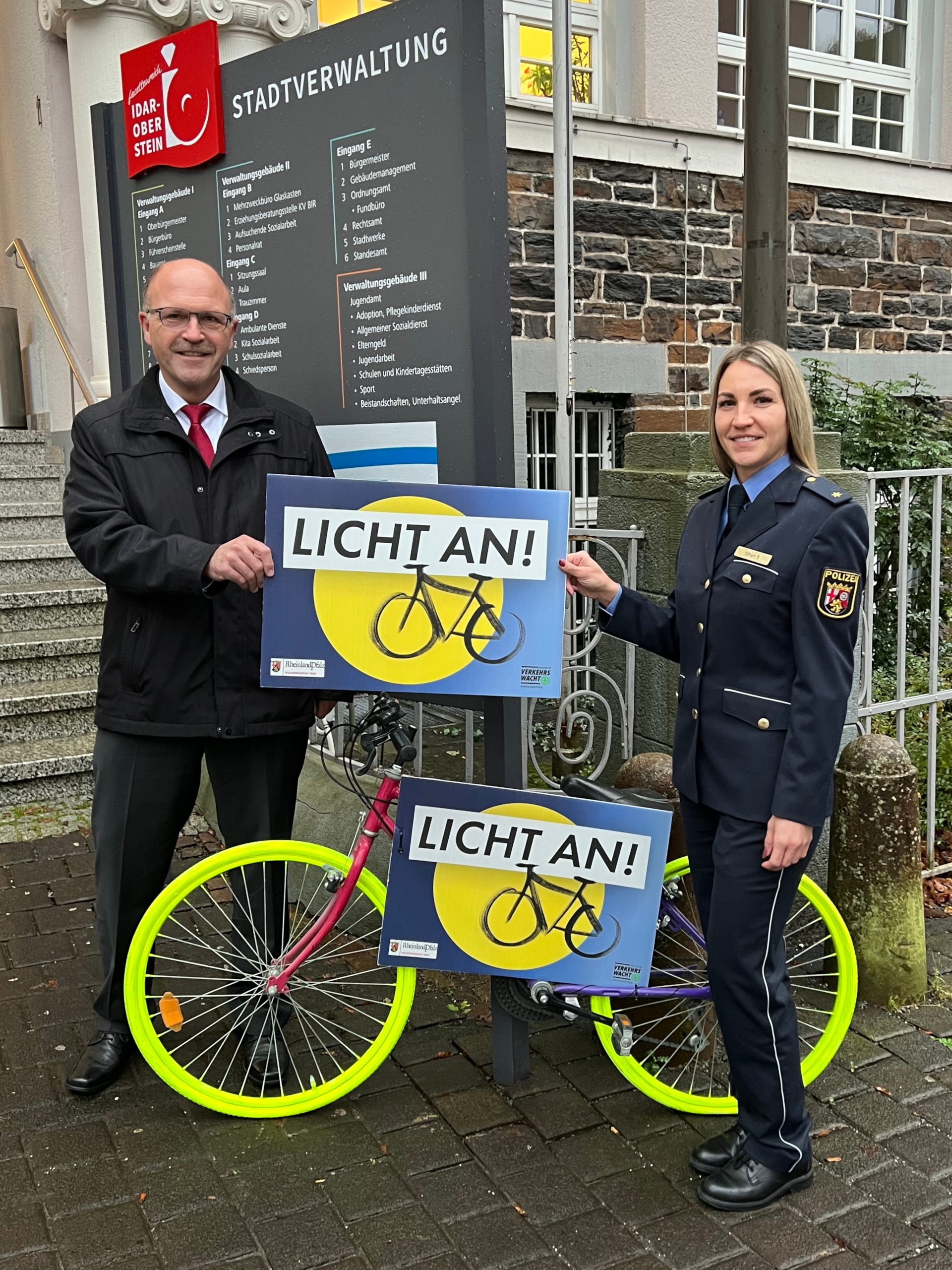 La photo montre le maire Marx et la conseillère de police Short. Ils se tiennent devant la mairie avec l'un des vélos tagués et tiennent ensemble un panneau sur lequel est écrit "Allumez les lumières".