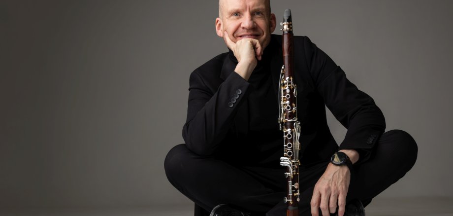 La photo montre le clarinettiste, en tenue noire, assis en tailleur sur le sol. Devant lui, son instrument est posé sur le sol.