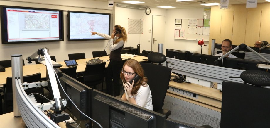 La photo montre une vue du centre d'alerte du BBK, une femme est assise devant un écran et parle au téléphone, une femme parle au téléphone et désigne un grand écran sur le mur, un homme regarde ses écrans.