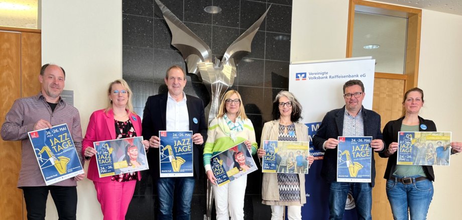 La photo montre sept personnes. Elles tiennent des affiches des Journées du jazz, se tiennent côte à côte devant une sculpture et regardent l'objectif.