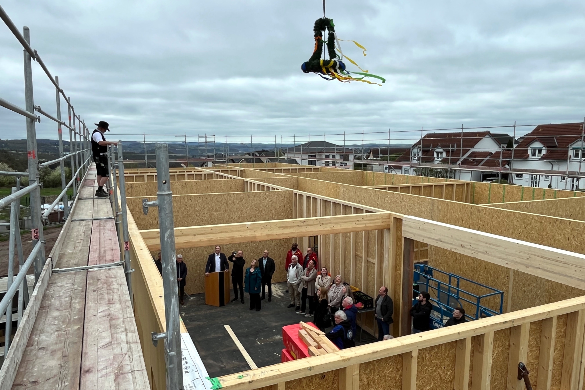 La photo montre une vue d'en haut du chantier. A gauche, sur l'échafaudage, le charpentier est en train de faire le bouquet final. En dessous, dans le bâtiment, les invités de la fête du bouquet final se tiennent debout et regardent le charpentier en haut.