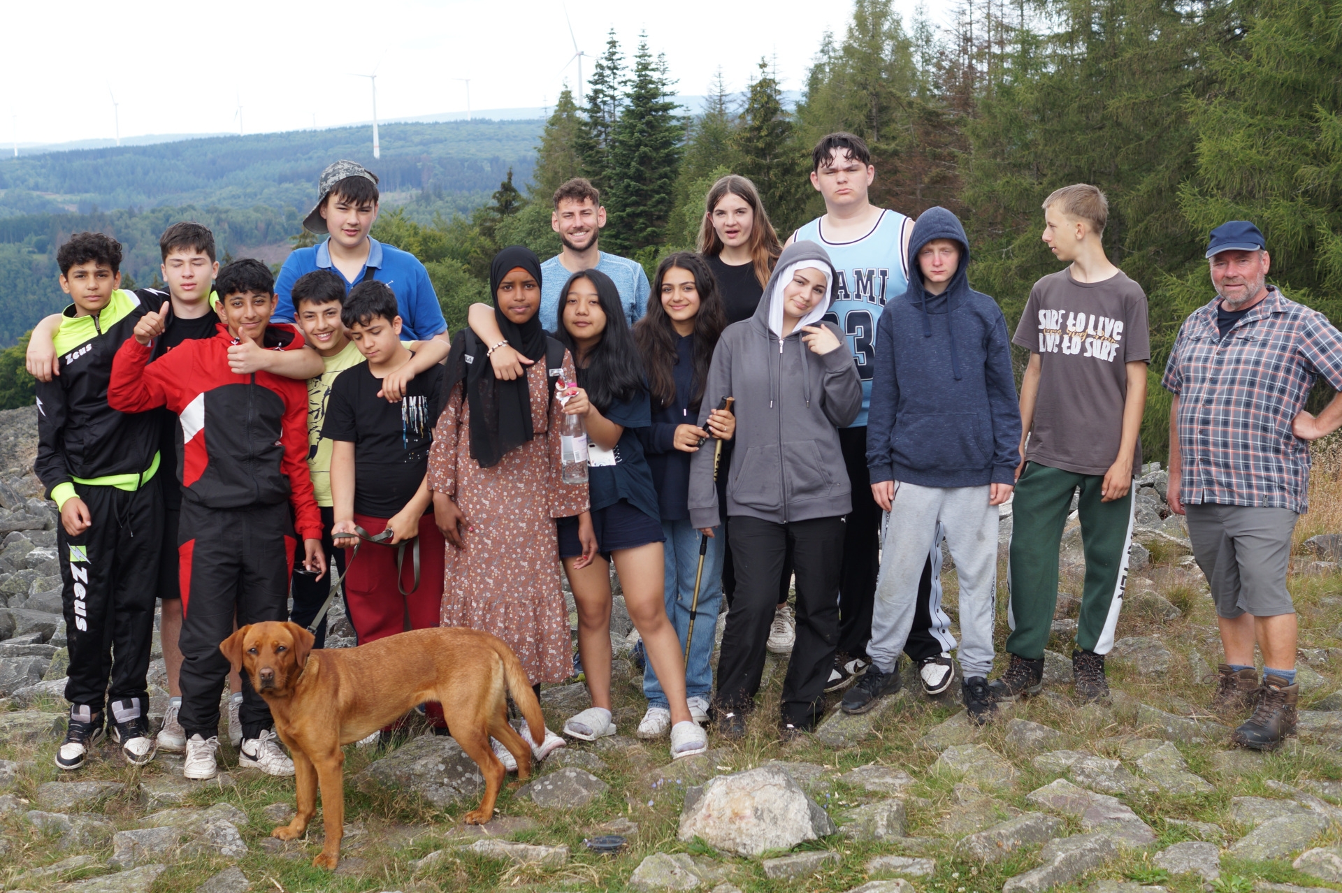 La photo montre les élèves et leurs accompagnateurs. Ils se tiennent en deux rangées côte à côte devant le dos d'une forêt et regardent la caméra. Un autre chien se tient devant le groupe.