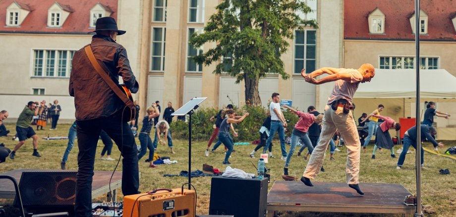 La photo montre au premier plan un guitariste sur scène. Un peu devant, on voit Sebastian Weber sur un élément de scène. Il montre aux participants une séquence de mouvements. Le tout se déroule en plein air.