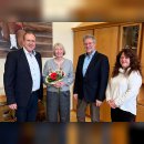 La photo montre les personnes mentionnées. Elles se tiennent côte à côte dans le bureau du maire. La personne qui fête son anniversaire tient un bouquet de fleurs dans ses mains.