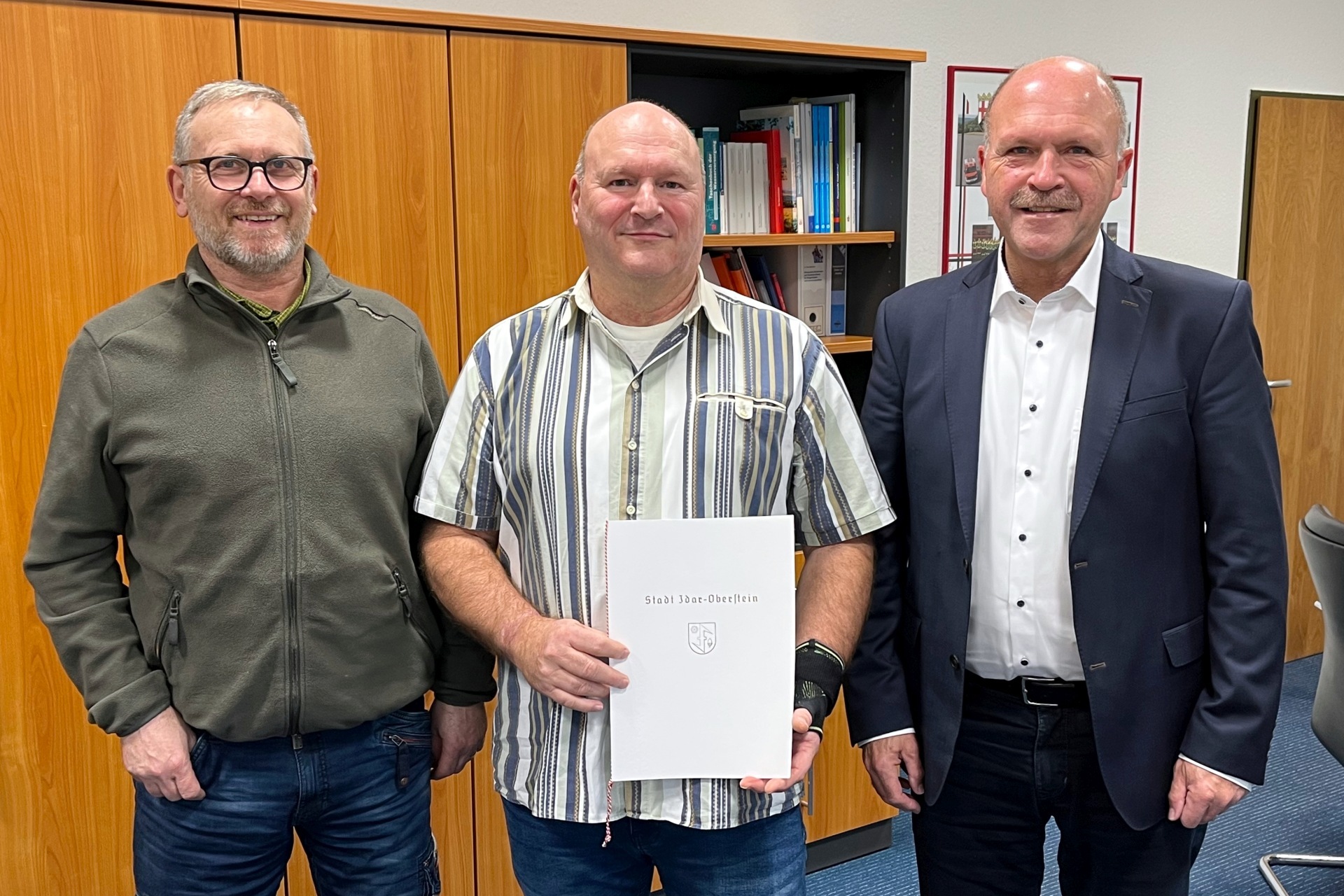 La photo montre trois personnes debout devant une armoire dans le bureau du maire Marx et regardant la caméra. M. Klintz se tient au milieu, tenant le certificat dans ses mains.