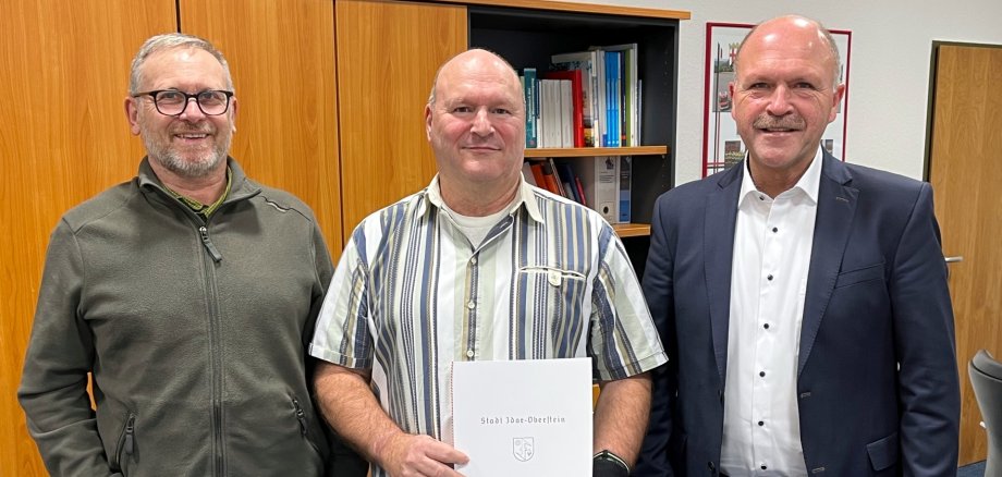 La photo montre trois personnes debout devant une armoire dans le bureau du maire Marx et regardant la caméra. M. Klintz se tient au milieu, tenant le certificat dans ses mains.