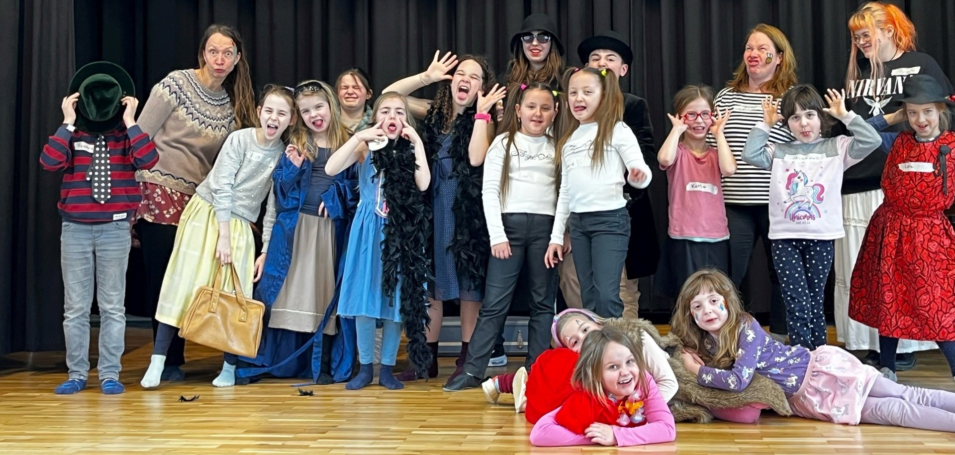 La photo montre les participants à la Journée du spectacle. Maquillés et costumés, ils se tiennent côte à côte sur la scène de l'aula Göttenbach et font des grimaces.