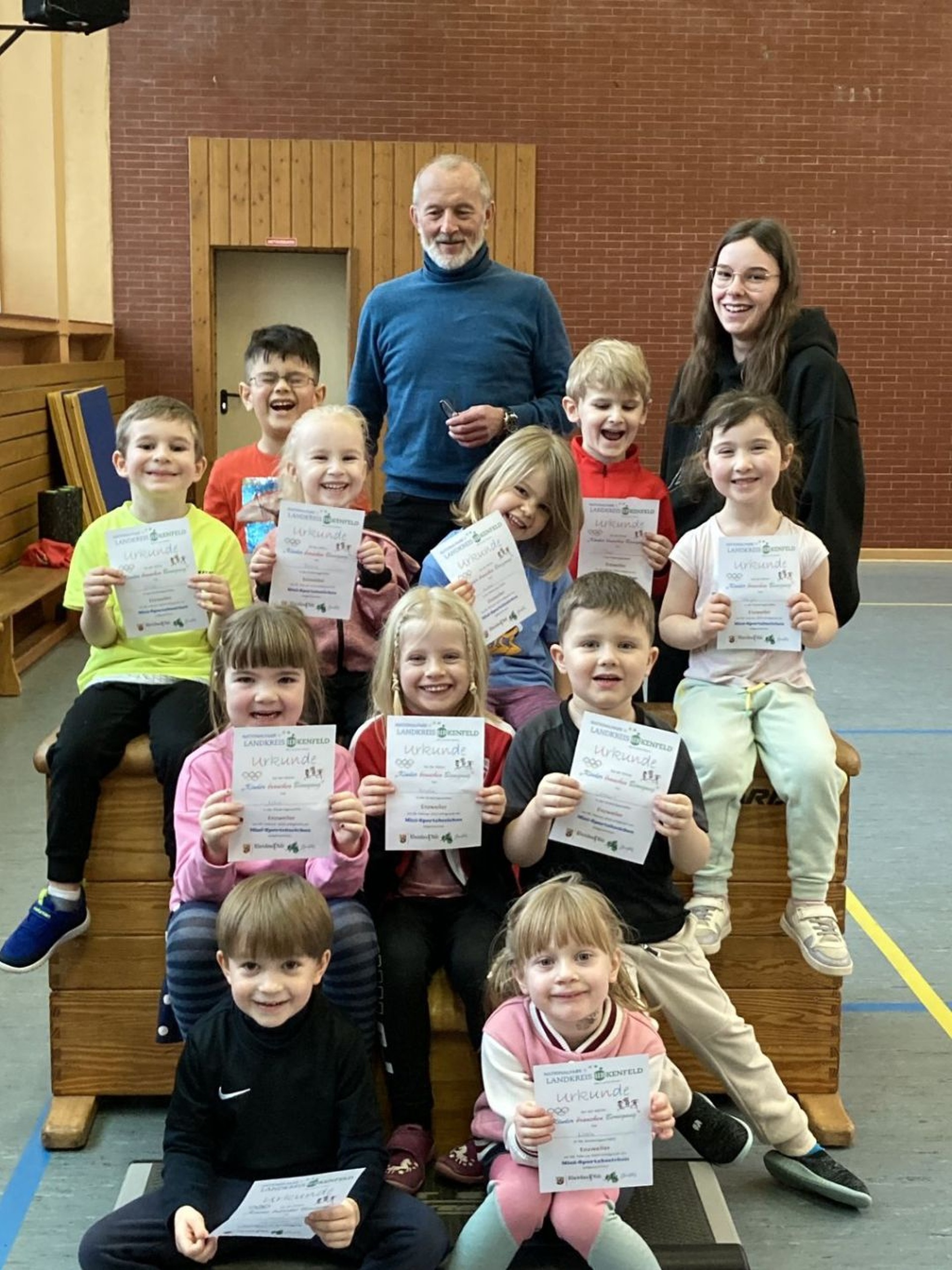 La photo montre onze enfants, Klaus Juchem et une éducatrice. Les enfants sont assis dans le gymnase devant et sur un caisson (l'équipement sportif) et tiennent des diplômes dans leurs mains. Les adultes se tiennent derrière eux.