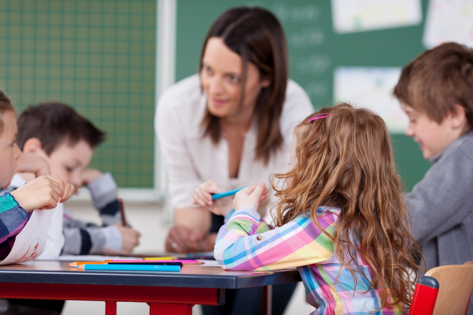 La photo montre une enseignante et plusieurs élèves dans une salle de classe. Les élèves sont assis à une table et l'enseignante se penche vers eux.